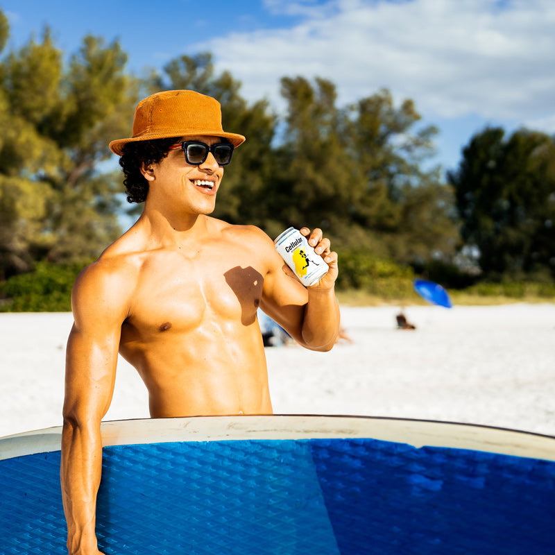 Man drinking Unsweetened Lemon Water flavor Cellular Hydration while holding surfboard on beach