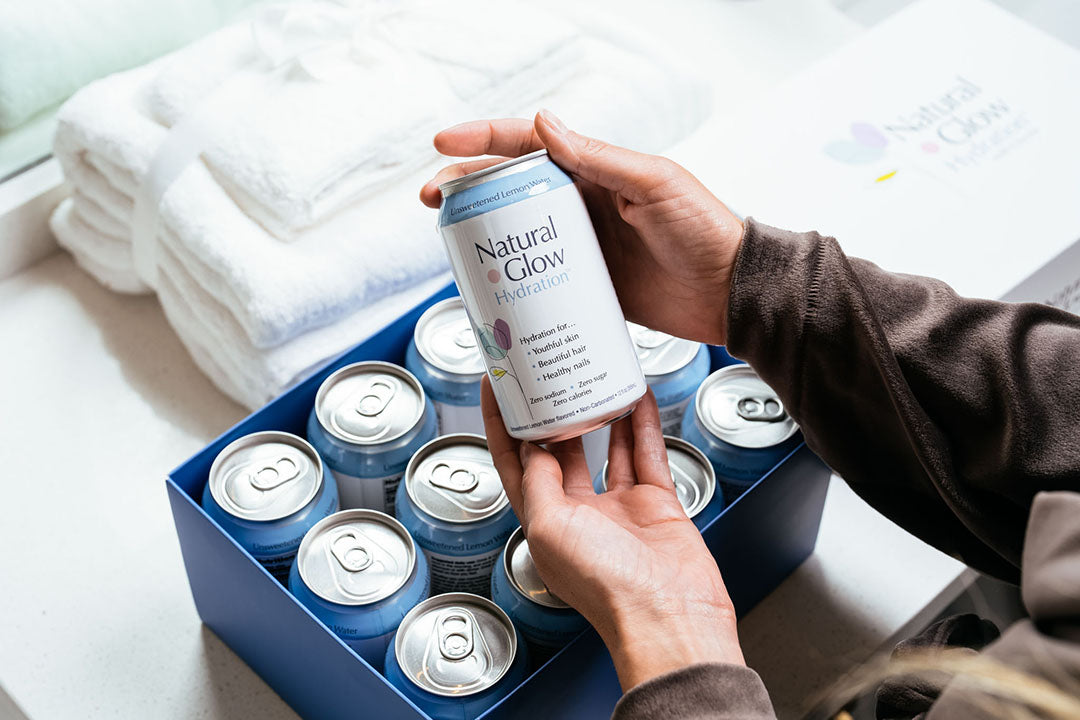 Woman holding can of Unsweetened Lemon Water flavor Natural Glow Hydration in front of 12-pack case on a bathroom counter