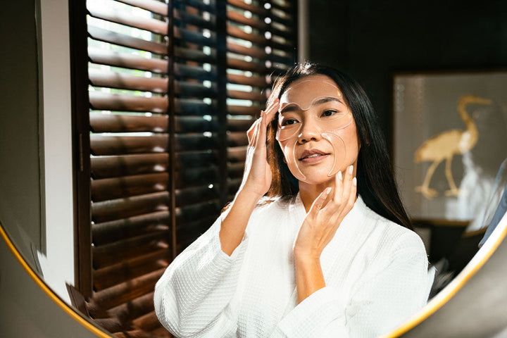 Asian woman applying Natural Glow Hydration Anti-Aging Facial Petals to her face wearing robe in bathroom mirror