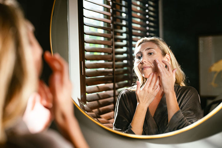 Young woman applying Natural Glow Hydration Anti-Aging Facial Petals to her face wearing robe in bathroom mirror