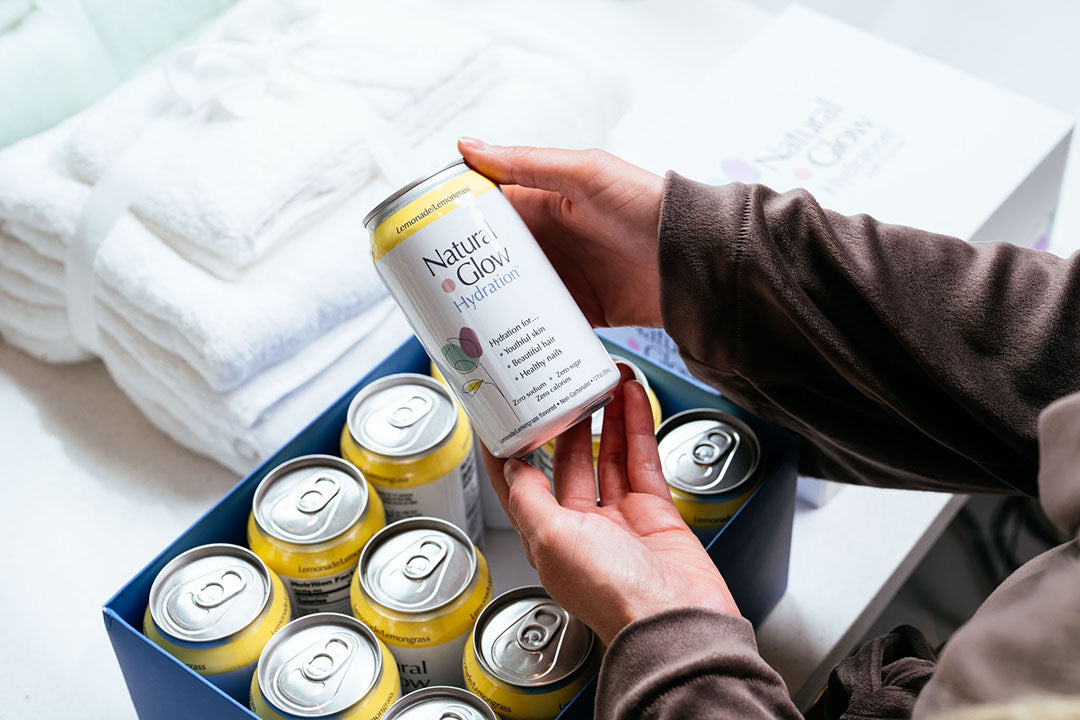 Woman holding can of Lemonade / Lemongrass flavor Natural Glow Hydration in front of 12-pack case on a bathroom counter