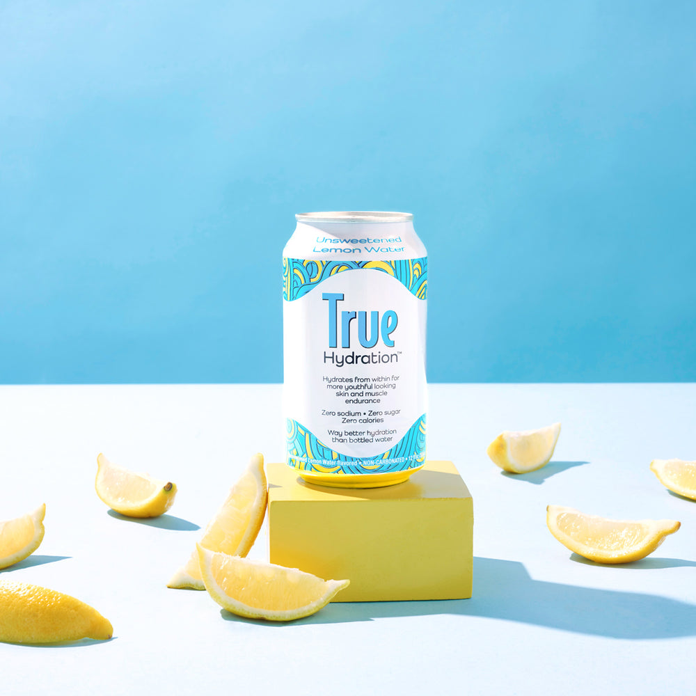 True Hydration Unsweetened Lemon Water Can on Yellow Pedestal with Lemons Blue Background