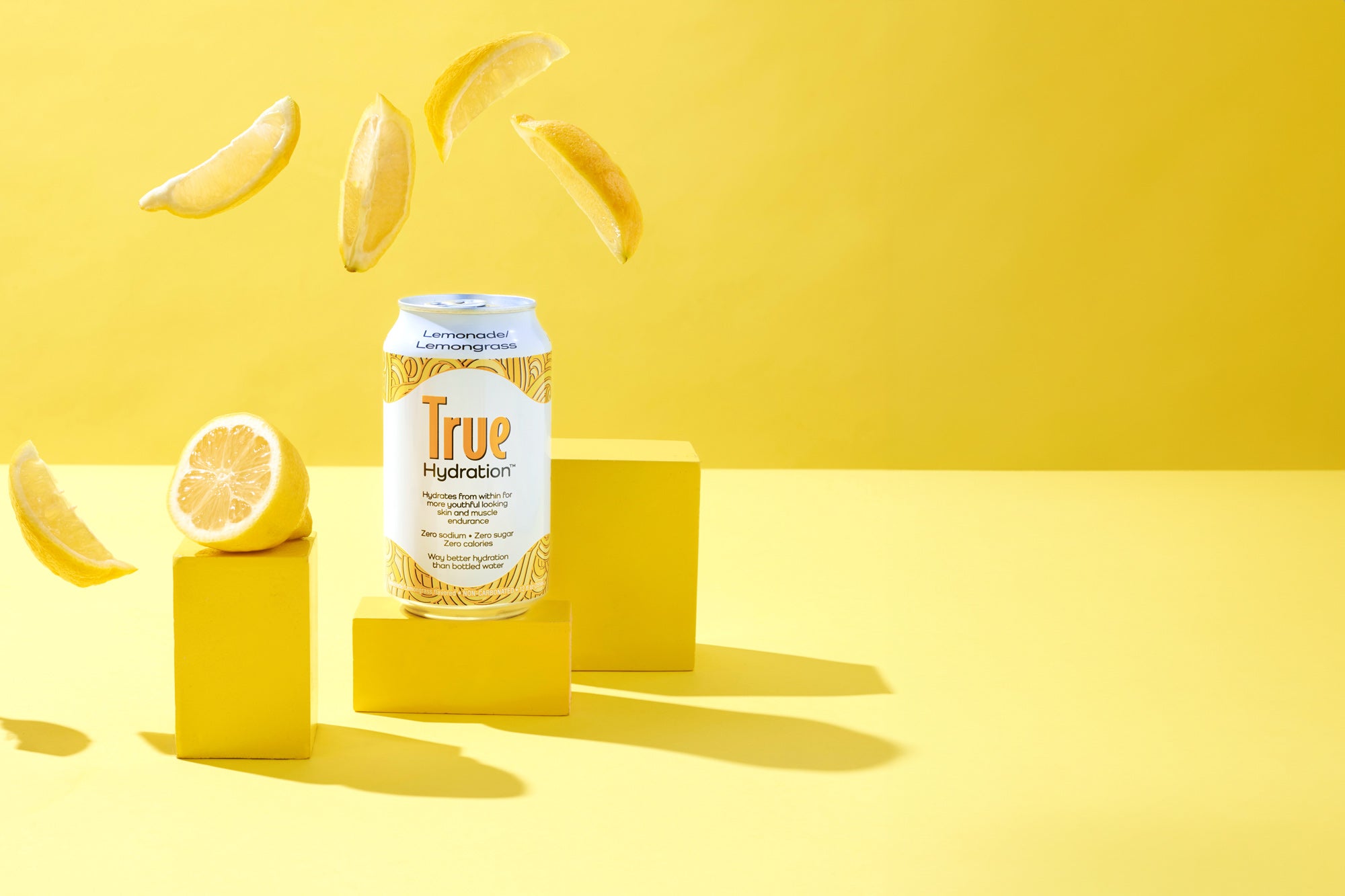 Can of Lemondade / Lemongrass flavor True Hydration on a yellow pedestal with lemon slices in the air on a yellow background